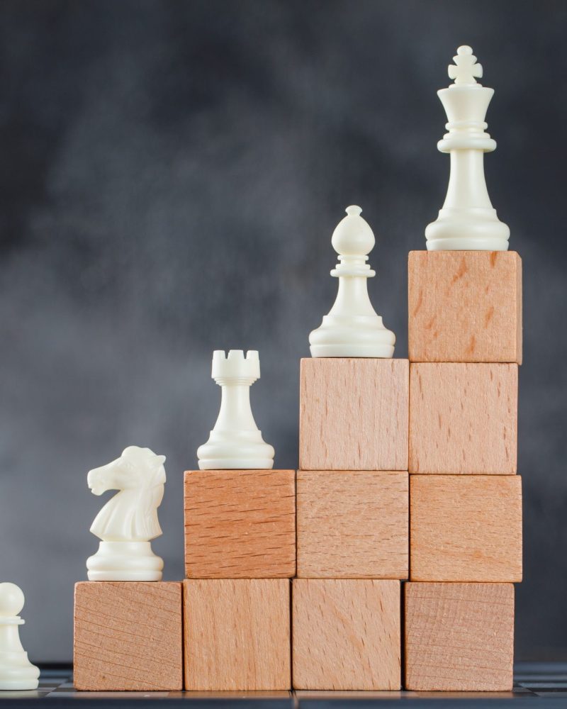 Business hierarchy concept with figures on pyramid of wooden blocks on foggy and chessboard background side view.